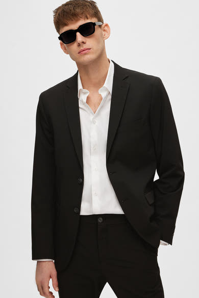 Zwarte blazer van SELECTED, gedragen over een open wit overhemd, met een zwarte zonnebril.