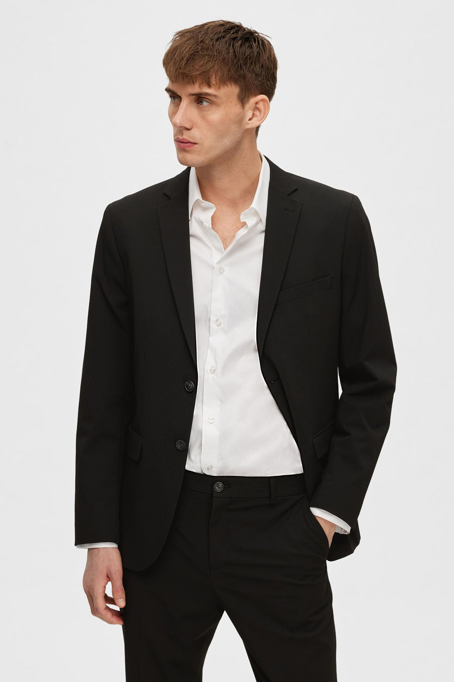 Zwarte blazer van SELECTED, gedragen over een open wit overhemd, bijpassende pantalon en hand in de zak.