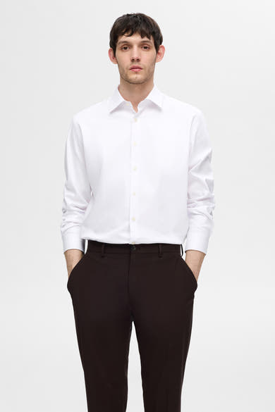 Chemise blanche à manches longues de SELECTED, portée avec un pantalon marron, mains dans les poches.
