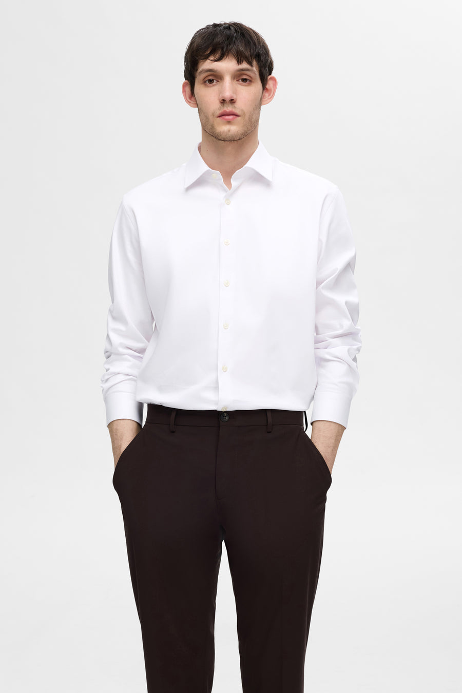 Chemise blanche à manches longues de SELECTED, portée avec un pantalon marron, mains dans les poches.
