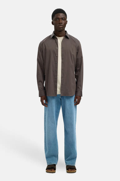 Chemise à manches longues marron de SELECTED, portée ouverte sur un t-shirt clair et un jean bleu décontracté.