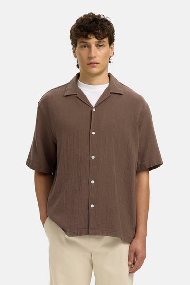 Chemise à manches courtes brune de SELECTED, en tissu texturé, col ouvert, boutons blancs, portée sur un t-shirt blanc avec un pantalon beige.