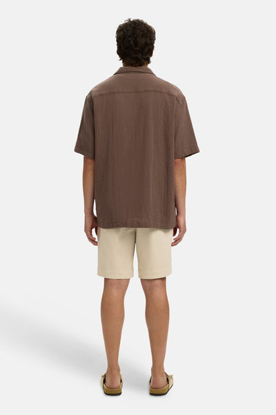Chemise à manches courtes brune de SELECTED, au tissu texturé et coupe ample, portée de dos avec un short beige et des sandales.