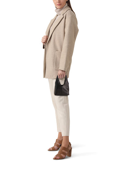 Zwarte handtas van MANGO, in de hand gedragen, met een beige jas, een witte broek en bruine sandalen met hak.