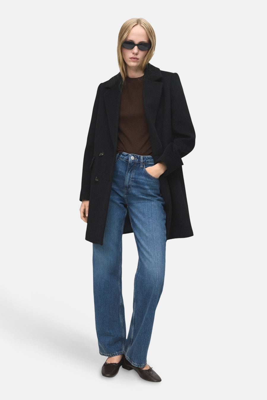 Veste longue noire de MANGO, portée avec un t-shirt marron, un jean bleu et des chaussures plates noires.