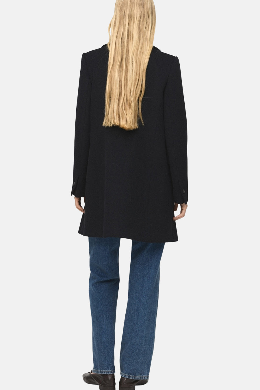 Veste longue noire par MANGO, avec une texture lisse et des boutons aux manches, portée avec un jean bleu et des chaussures plates.
