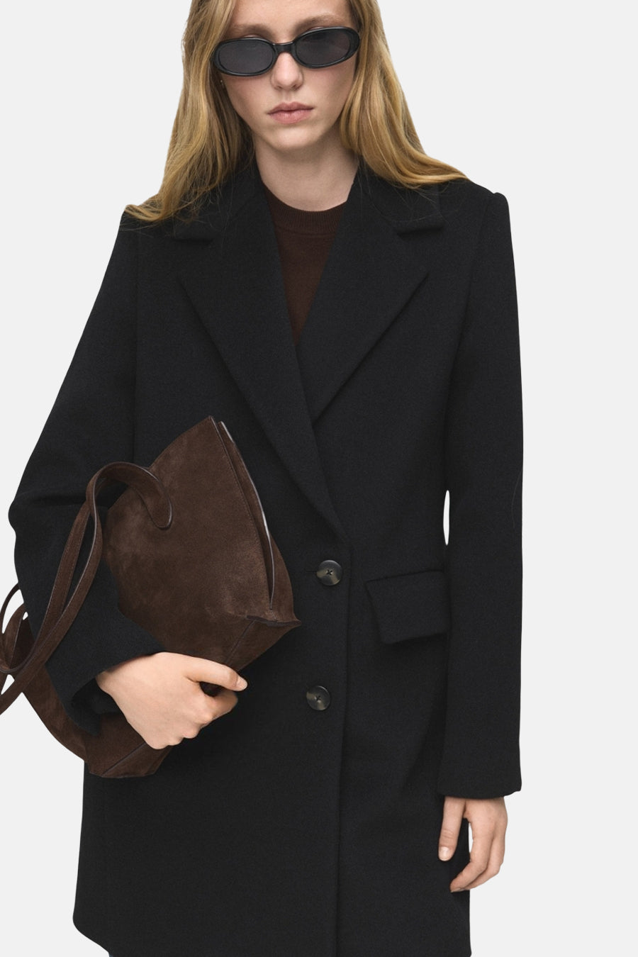 Veste longue noire de MANGO, avec un col à revers, portée sur un pull marron et tenant un sac à main marron.