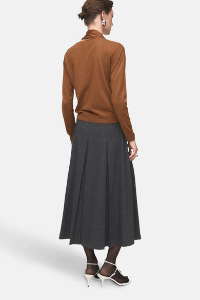 Pull à col rond brun de MANGO, porté avec une jupe midi plissée anthracite et des chaussures blanches à talons aiguilles.