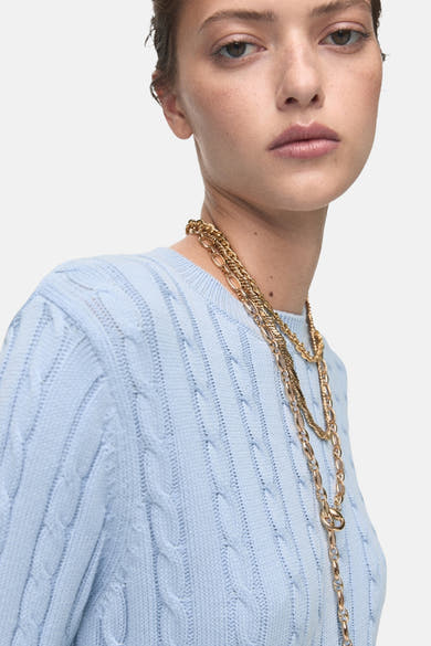 Pull bleu à col rond de MANGO, en tricot torsadé, porté avec un collier doré à plusieurs rangs.
