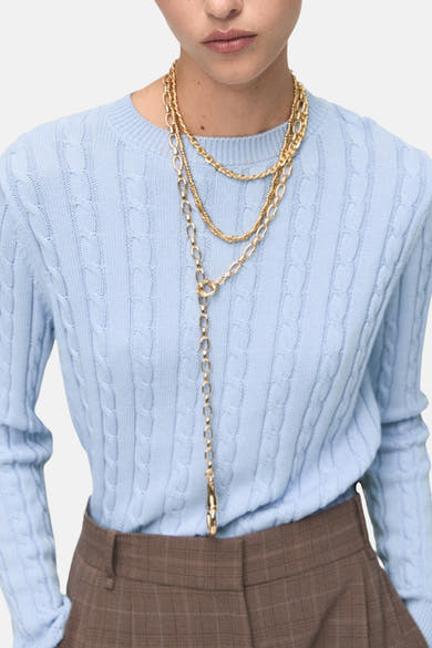 Pull à col rond bleu de MANGO, en maille torsadée, porté avec un pantalon à carreaux et des colliers dorés.
