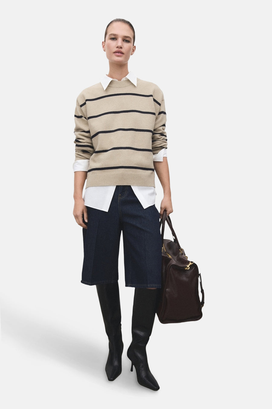Pull à col rond beige rayé de MANGO, porté sur une chemise blanche, avec un short en jean et des bottes noires.
