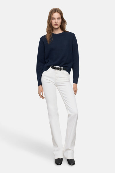 Pull à col rond bleu de MANGO, porté avec un pantalon blanc taille haute et une ceinture noire.