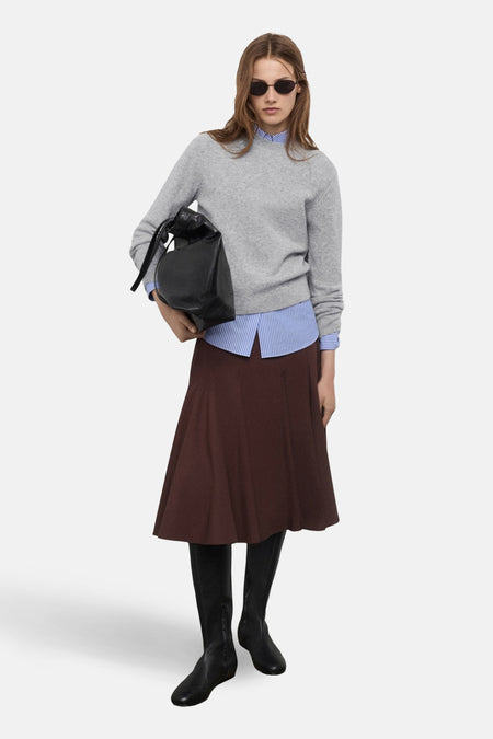 Pull à col rond gris de MANGO, porté avec une jupe marron, une chemise rayée bleue et des bottes noires.