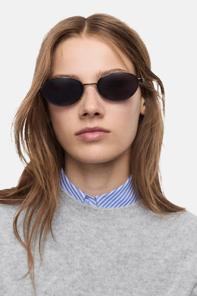 Pull à col rond gris de MANGO, porté sur une chemise rayée, avec des lunettes de soleil ovales.
