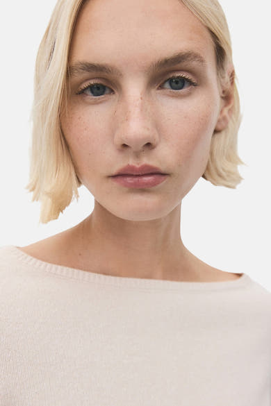 Pull écru à col rond de MANGO, porté par un modèle aux cheveux blonds, avec des taches de rousseur.
