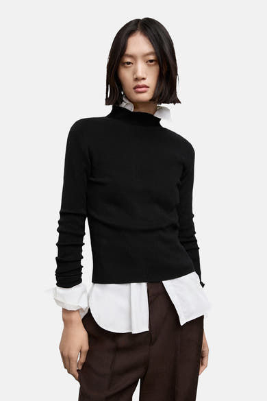 Pull à col rond noir de MANGO, porté sur une chemise blanche, avec un pantalon marron.