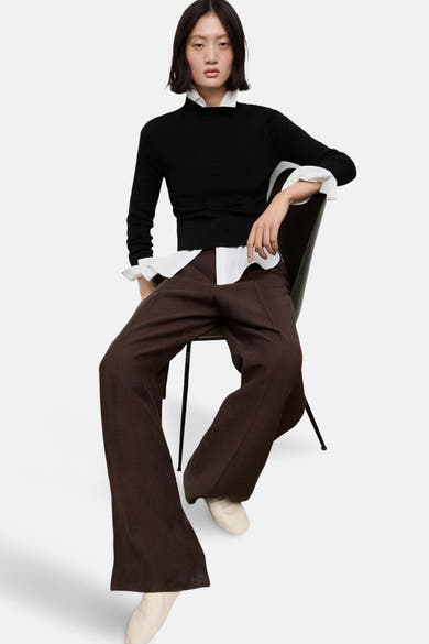 Pull à col rond noir de MANGO, porté avec une chemise blanche et un pantalon marron ample.