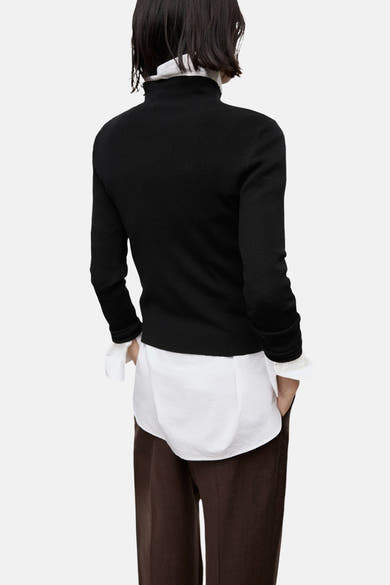 Pull à col rond noir de MANGO, porté avec une chemise blanche à col montant et un pantalon marron.