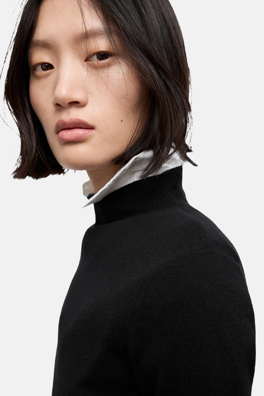 Pull à col rond noir de MANGO, porté avec une chemise blanche à col visible.
