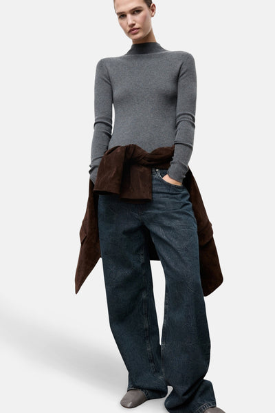 Pull à col rond gris de MANGO, porté avec un pantalon ample en denim et une veste marron nouée à la taille.
