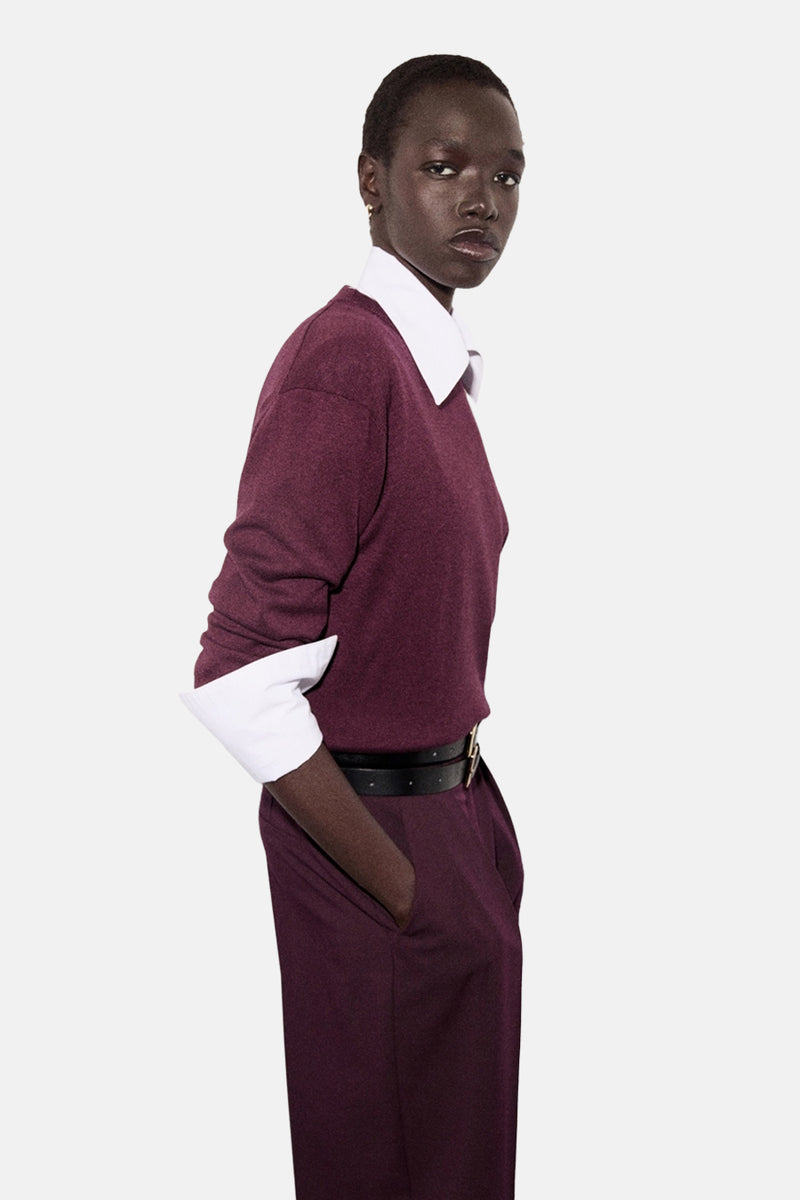 Pull à col rond bordeaux de MANGO, porté avec un chemisier blanc et une ceinture noire.
