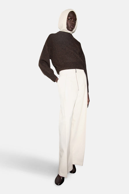 Pull à col rond marron de MANGO, porté avec un pantalon blanc à taille haute et fermeture éclair.
