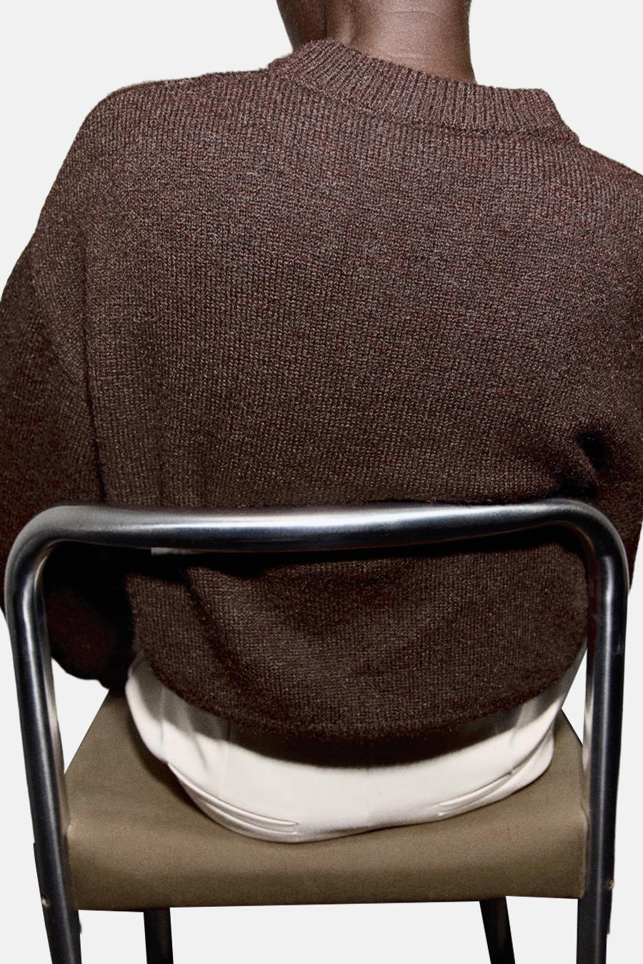 Pull à col rond brun de MANGO, vu de dos, porté avec un pantalon blanc sur une chaise en métal.
