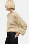 Pull à col rond beige de MANGO, en maille ajourée, porté avec un pantalon noir taille haute.
