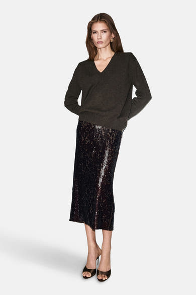 Pull à col en V brun de MANGO, porté avec une jupe midi à paillettes et des talons ouverts.
