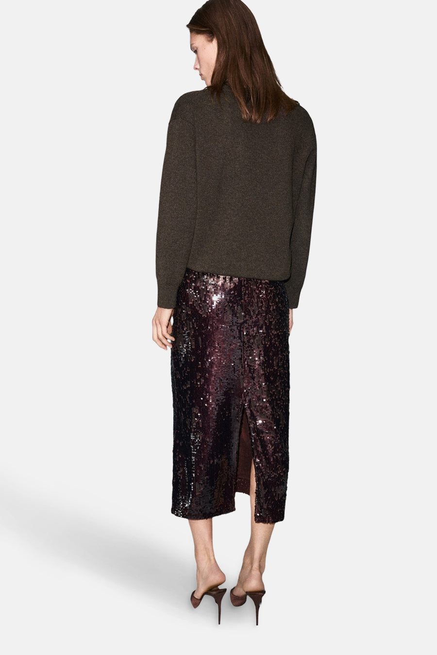 Pull à col en V marron de MANGO, porté avec une jupe à paillettes bordeaux et des mules à talons.