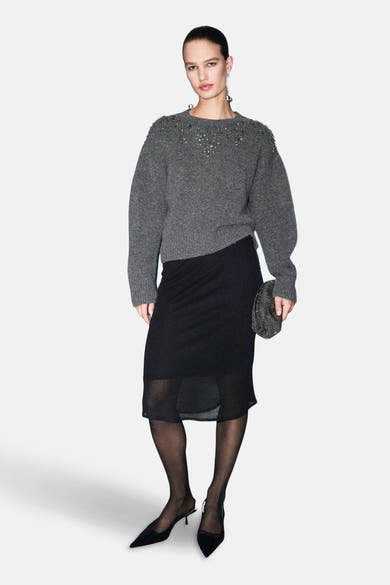 Pull à col rond gris de MANGO, orné de détails décoratifs aux épaules, porté avec une jupe noire et des collants.
