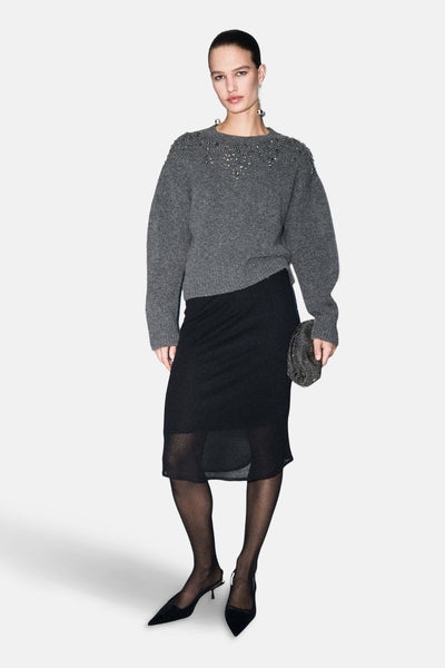 Grijze rondehals trui van MANGO, versierd met decoratieve details op de schouders, gedragen met een zwarte rok en panty.