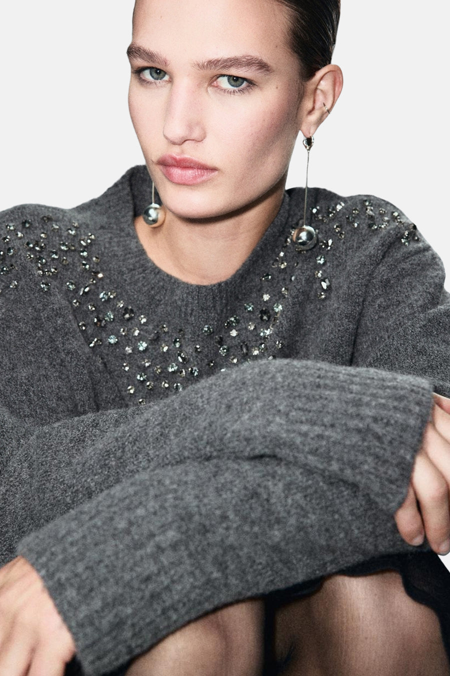Pull à col rond gris de MANGO, orné de pierres décoratives sur les épaules et porté avec des boucles d'oreilles argentées.
