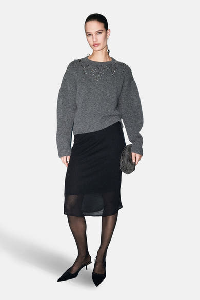 Pull à col rond gris par MANGO, orné de détails décoratifs, porté avec une jupe noire et des escarpins.
