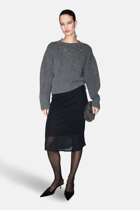Pull à col rond gris par MANGO, orné de détails décoratifs, porté avec une jupe noire et des escarpins.
