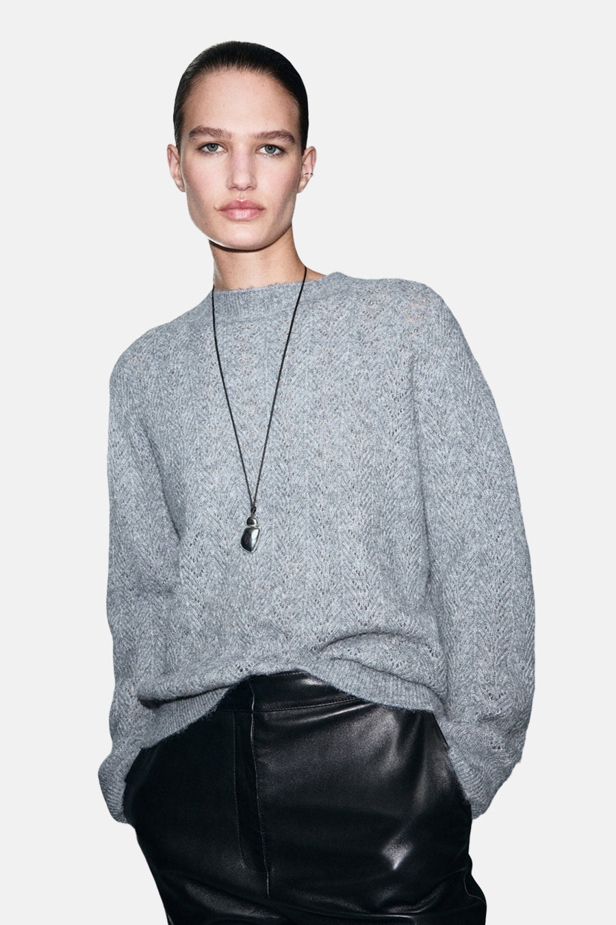 Pull à col rond bleu de MANGO, avec motif à chevrons, porté avec un pantalon en cuir noir et un collier pendentif.
