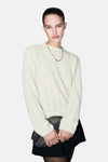 Pull à col rond écru de MANGO, à motif ajouré, porté avec une jupe noire et un sac à main brillant.
