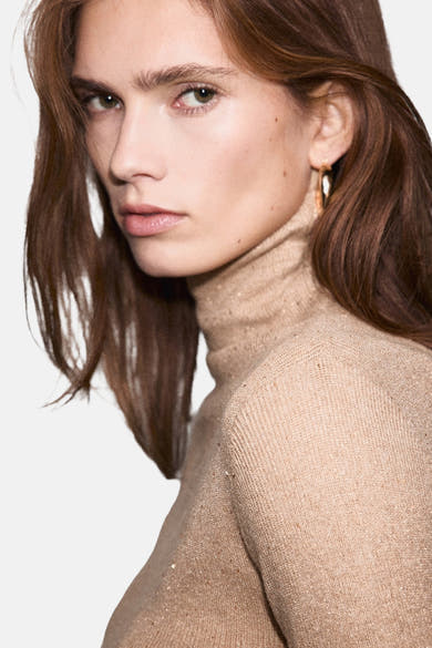 Pull à col roulé beige de MANGO, en maille fine brillante et portés avec des boucles d'oreilles dorées.