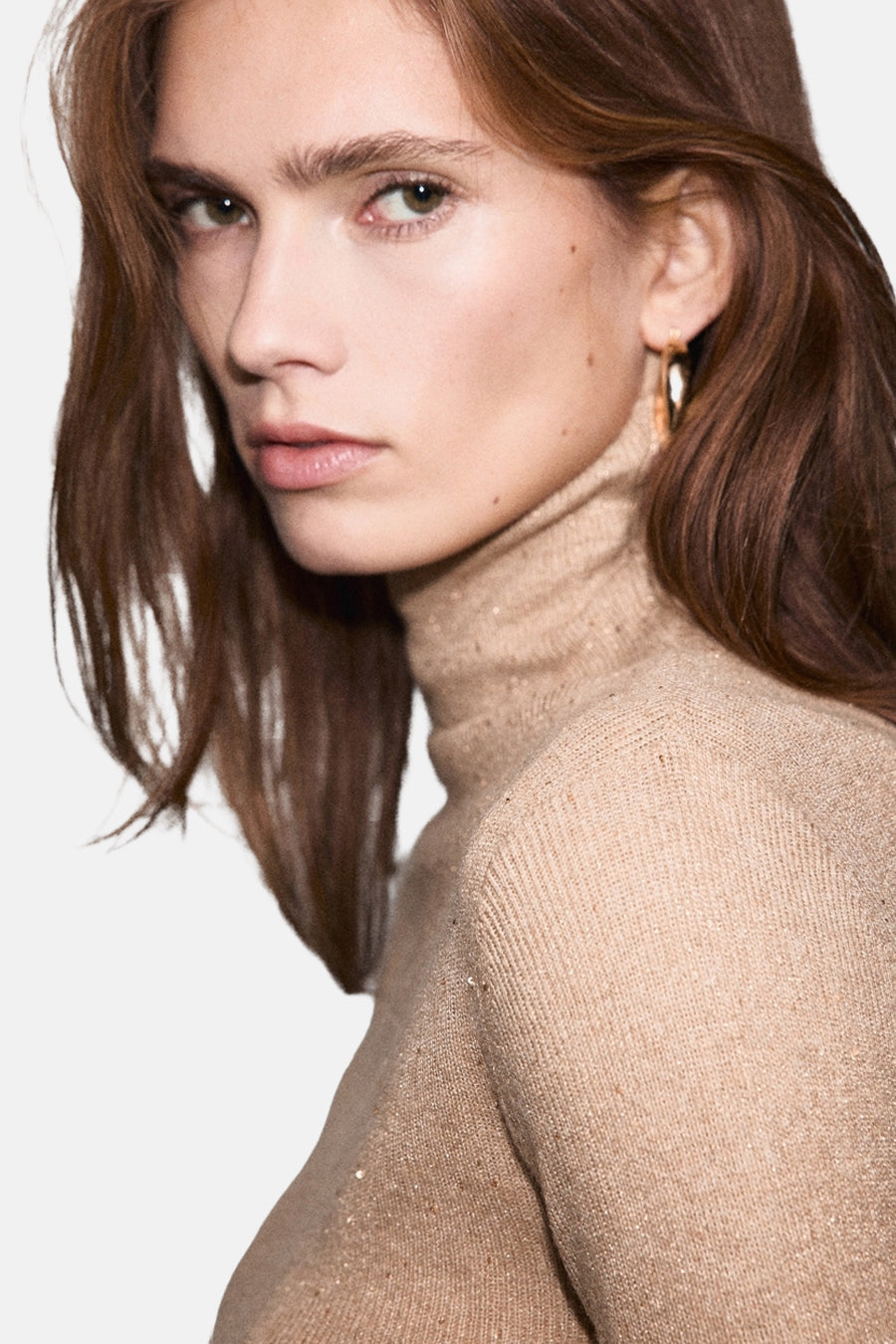 Pull à col roulé beige de MANGO, en maille fine brillante et portés avec des boucles d'oreilles dorées.