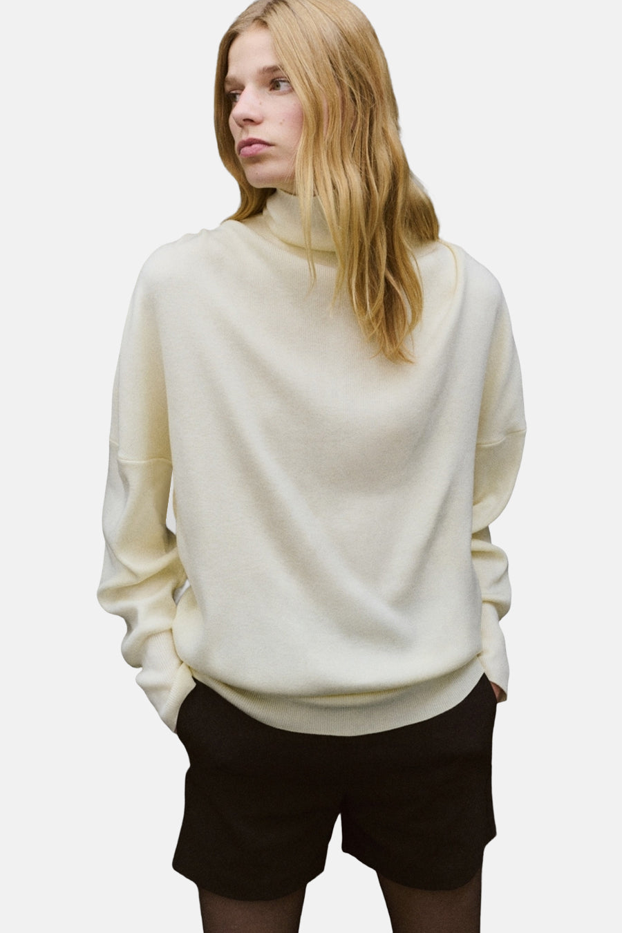 Pull à col roulé écru de MANGO, porté avec un short noir opaque et des collants noirs.