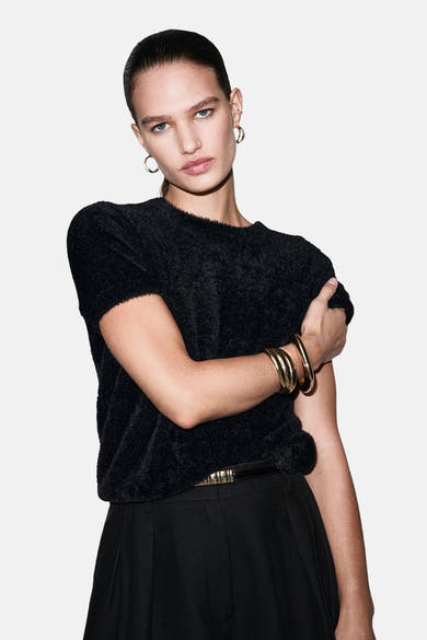 Pull à col rond noir de MANGO, avec des bracelets dorés et une ceinture noire.