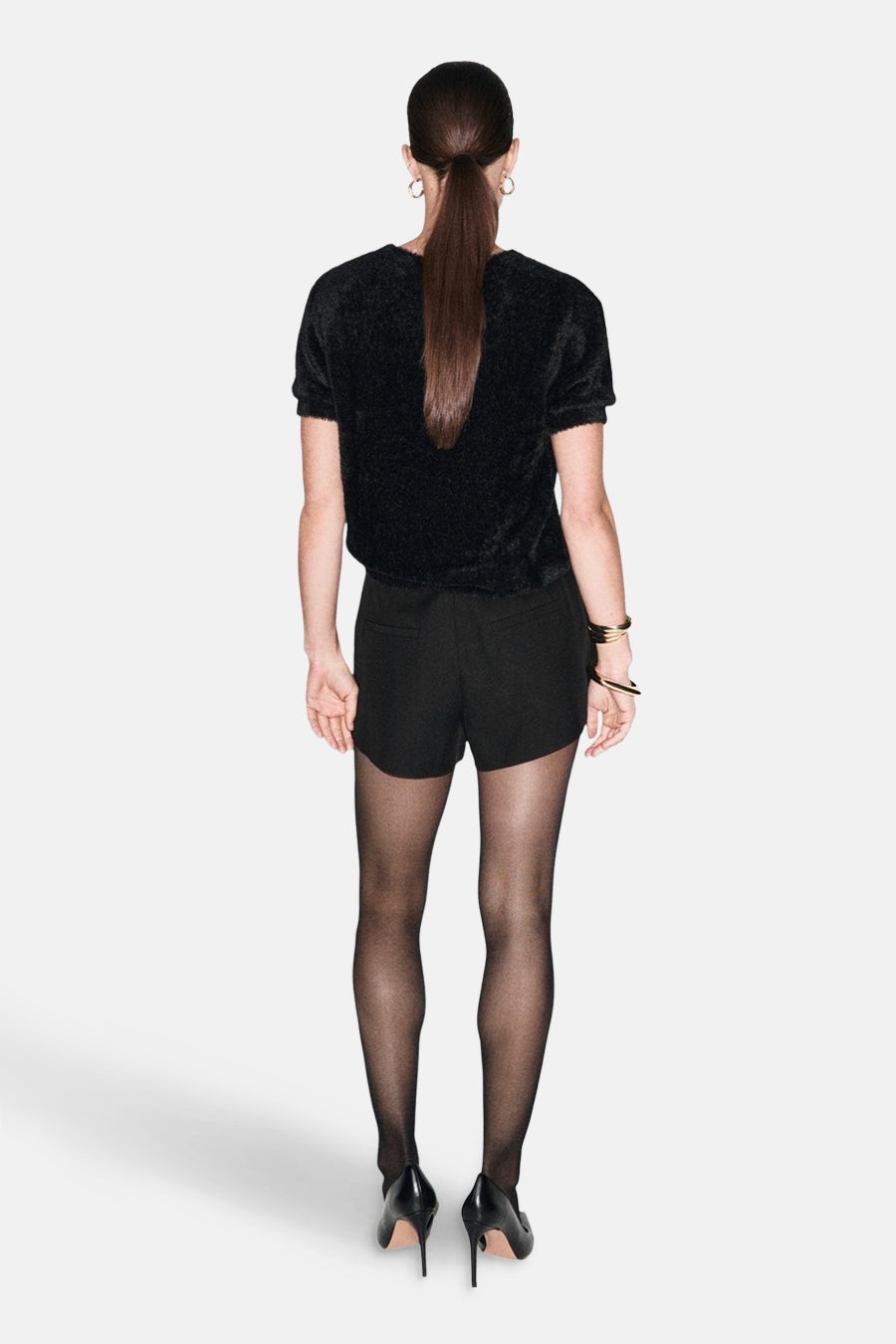 Pull à col rond noir de MANGO, porté avec un short noir, des collants noirs et des escarpins noirs.