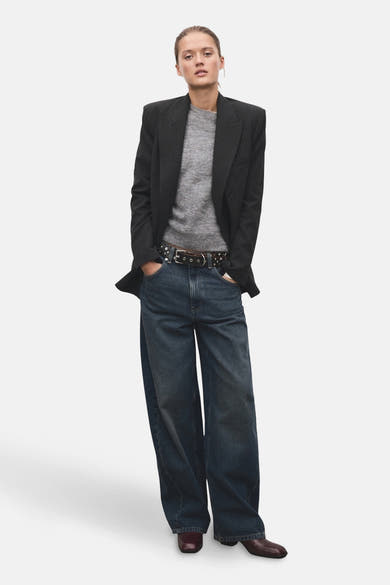 Pull à col rond gris de MANGO, porté avec un blazer anthracite, un jean large et une ceinture cloutée.