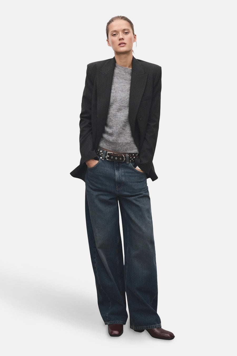Pull à col rond gris de MANGO, porté avec un blazer anthracite, un jean large et une ceinture cloutée.