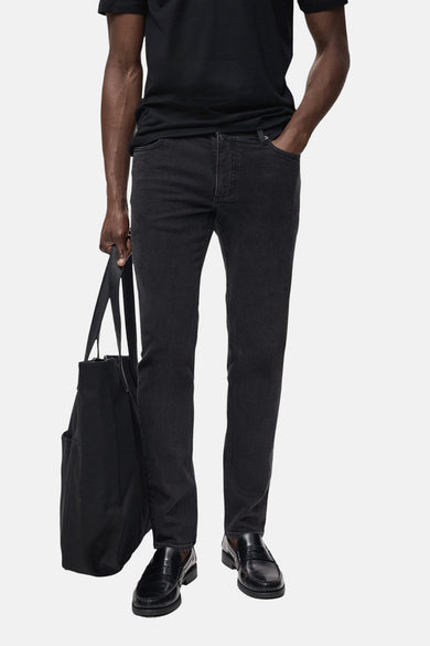 Donkergrijze spijkerbroek van MANGO, gedragen met een zwart T-shirt en leren loafers, met een zwarte canvas tas.