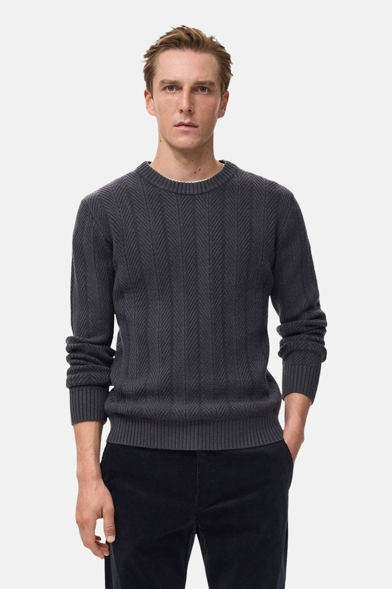 Pull à col rond gris de MANGO, avec un motif à chevrons texturé, porté avec un pantalon en velours côtelé.