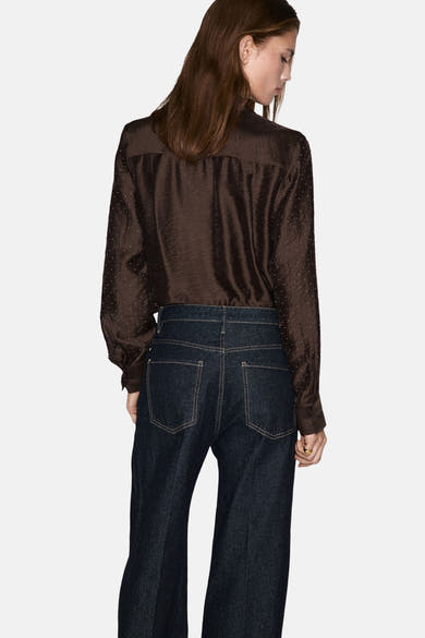 Chemisier marron à manches longues de MANGO, avec motif à pois et détails plissés, porté avec un jean bleu foncé.
