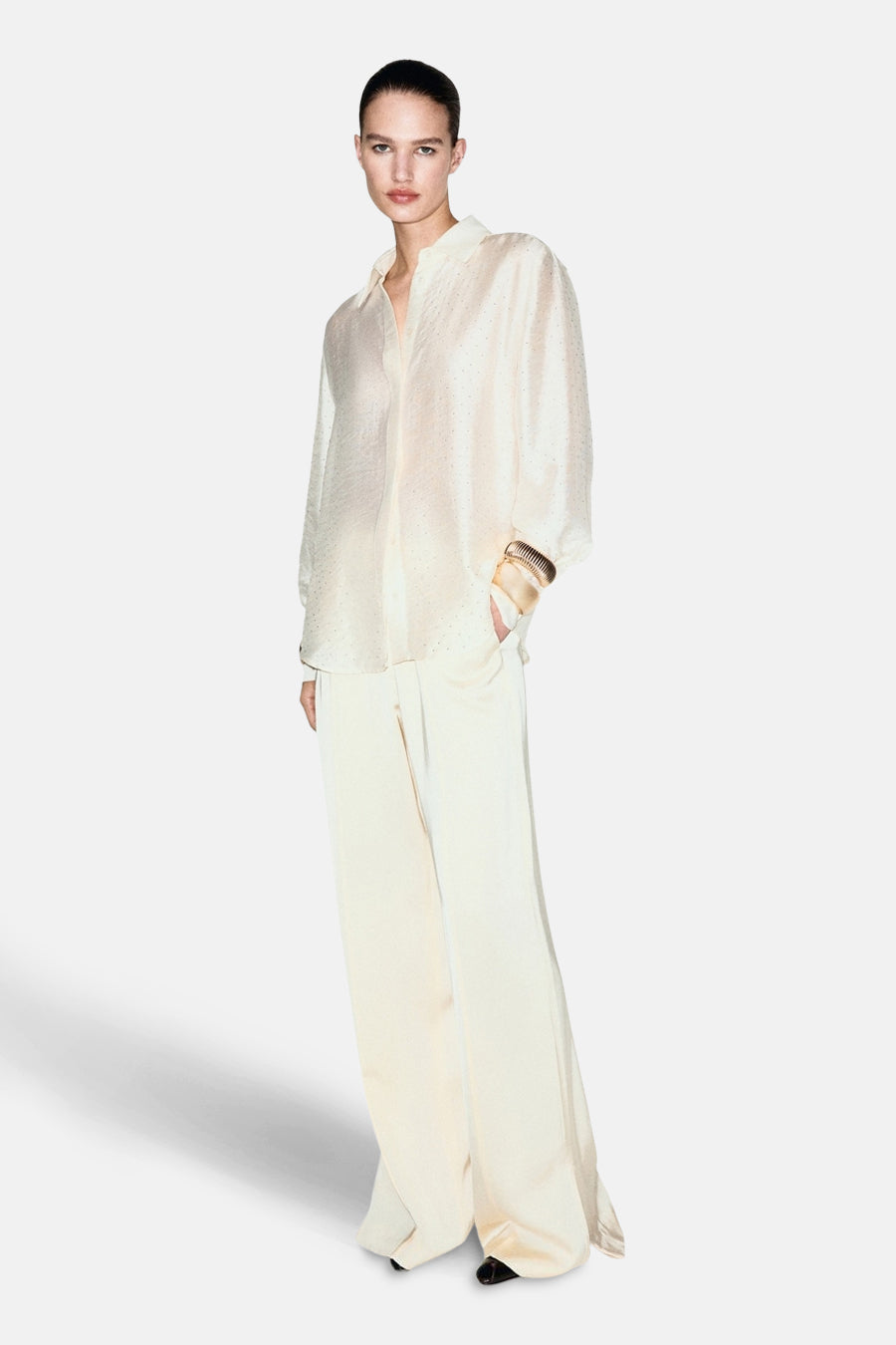 Chemise à manches longues écru de MANGO, avec une texture subtile, portée avec un pantalon fluide assorti et des manchettes dorées.
