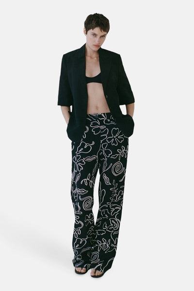 Pantalon noir à motif fleuri de MANGO, porté avec une brassière noire et un blazer noir.