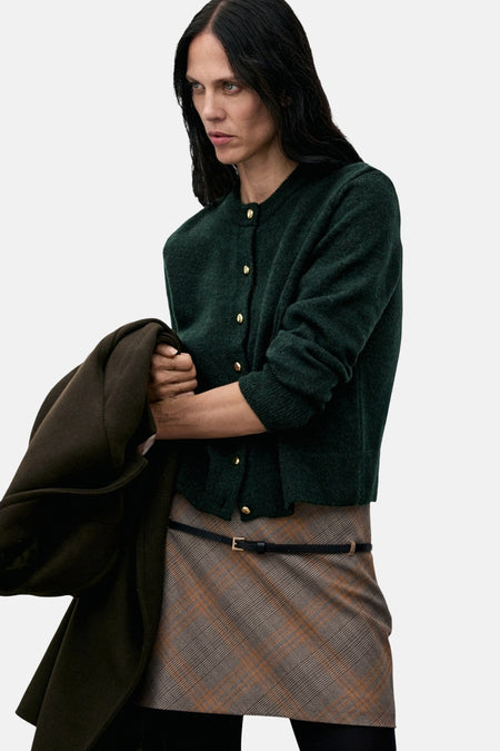 Groene cardigan van MANGO, met een zachte textuur en lange mouwen, gedragen met een geruite rok en zwarte panty.
